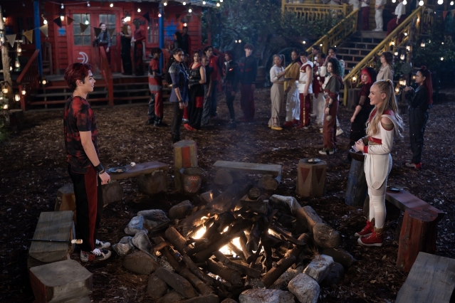 A young man and woman face each other across a campfire, with a large group gathered outside rustic cabins under string lights.